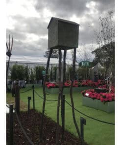 Birdhouse sculpture on tall wooden legs in The Kildare Gallery garden, surrounded by colourful flowers and landscaped greenery.