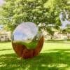 Reflective metallic sphere art installation in Kildare Garden, surrounded by lush greenery and trees.