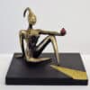 Glossy bronze abstract sculpture of a seated female figure with a long neck and stylized head, holding a small red object, on black base.