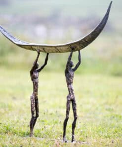 Bronze sculpture of two figures holding a crescent-shaped artwork, set outdoors in a grassy area.