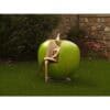 Vintage garden sculpture with a metallic figure sitting inside a large, green apple-shaped chair, surrounded by greenery and stone wall.