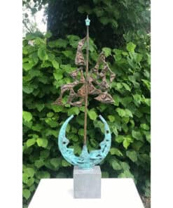 Rustic bronze and copper abstract sculpture featuring geometric shapes, mounted on a cubic stone plinth, set against lush green foliage.