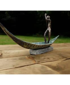 A weathered metal sculpture of a person on a carved canoe, placed on a wooden surface outdoors.