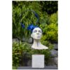 Vibrant blue artistic sculpture of a woman's bust with an abstract headpiece, displayed outdoors amidst lush green foliage.
