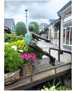 Vibrant outdoor sculpture of a girl with outstretched arms in Kildare town, surrounded by flowers and retail shops.